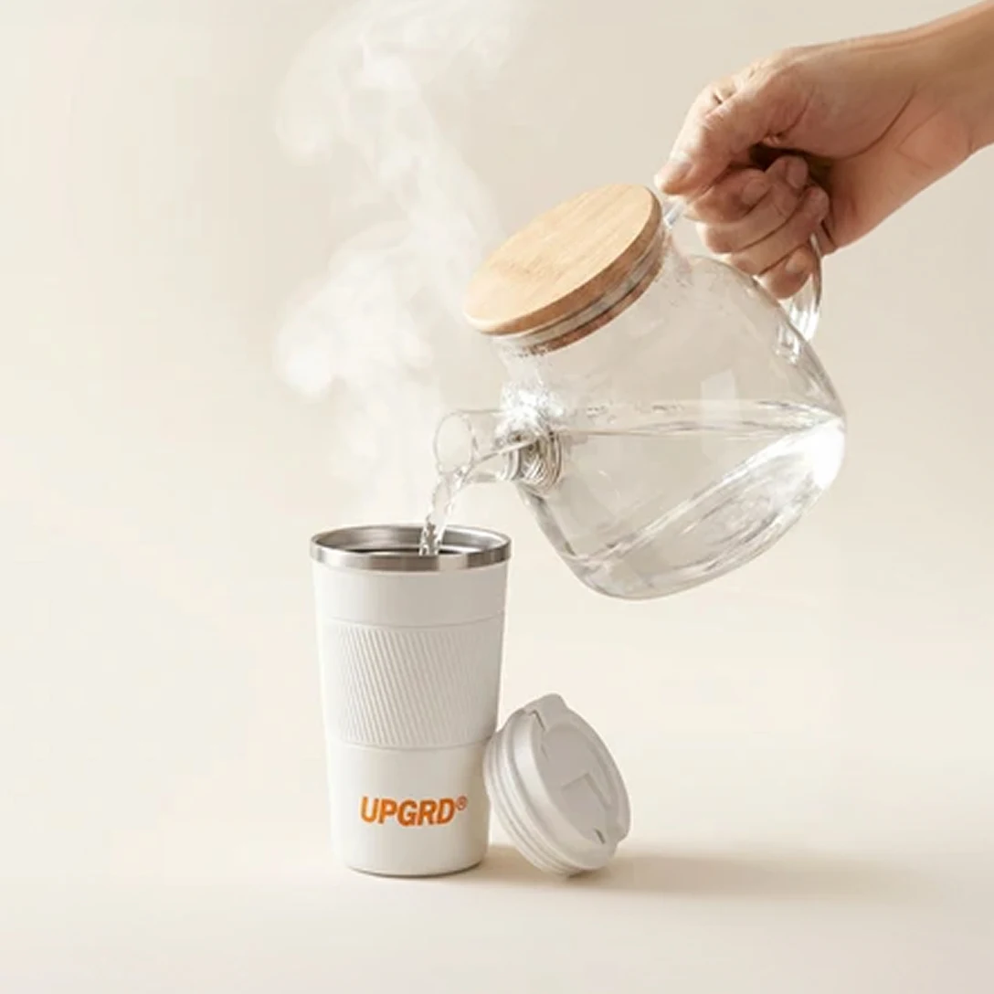 A hand pours steaming water from a glass pot into a white travel mug with an orange logo.