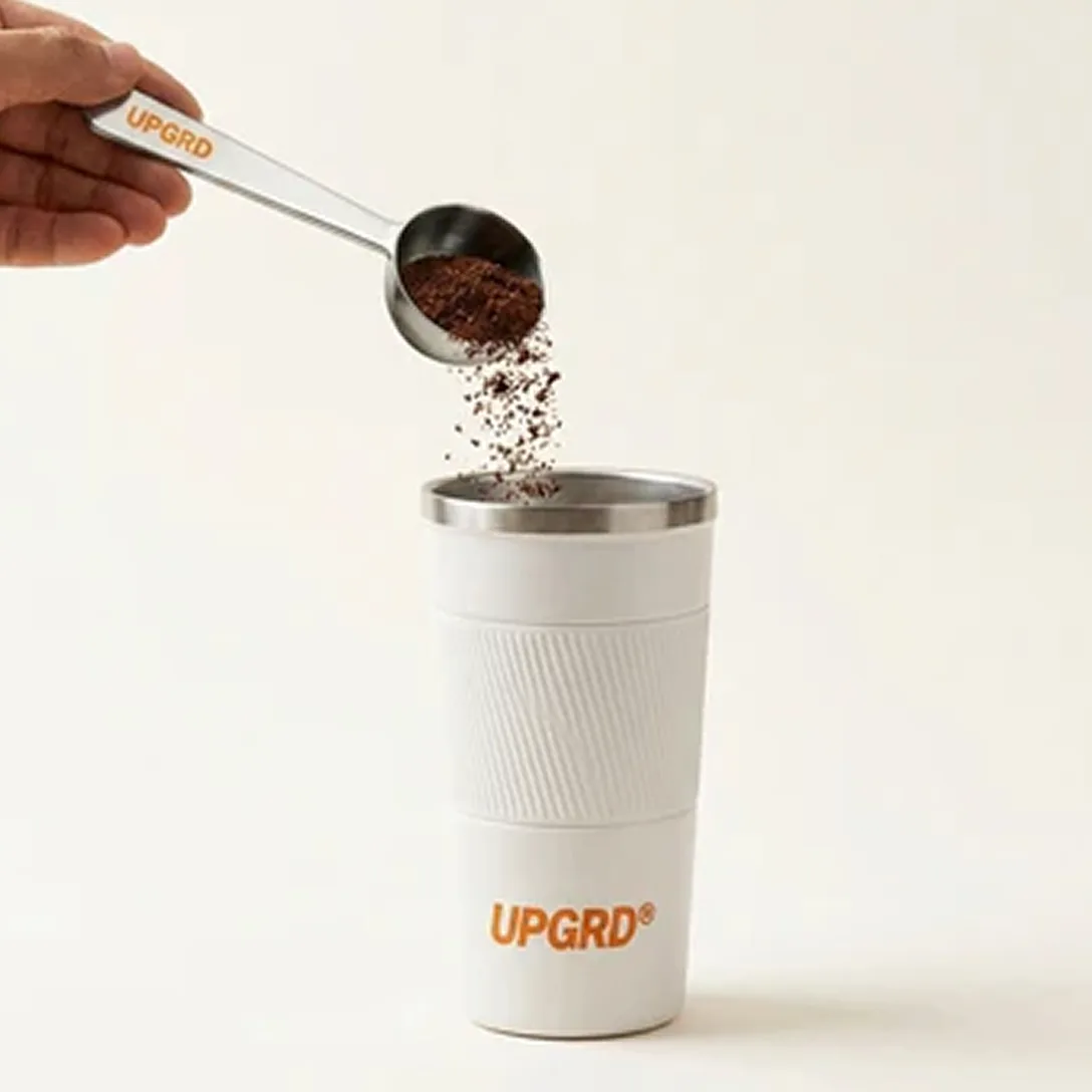 A hand pours coffee grounds from a metal scoop into a white UPGRD travel mug.