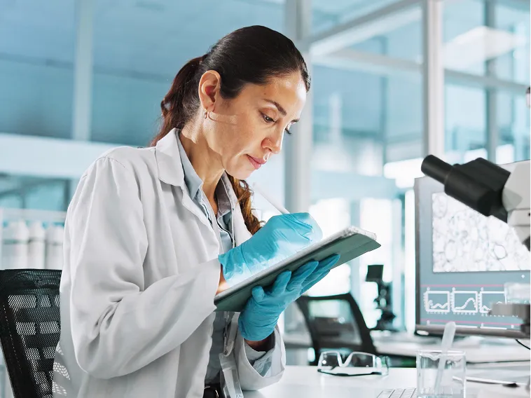A female scientist in a lab coat and gloves writes on a tablet in a laboratory setting.