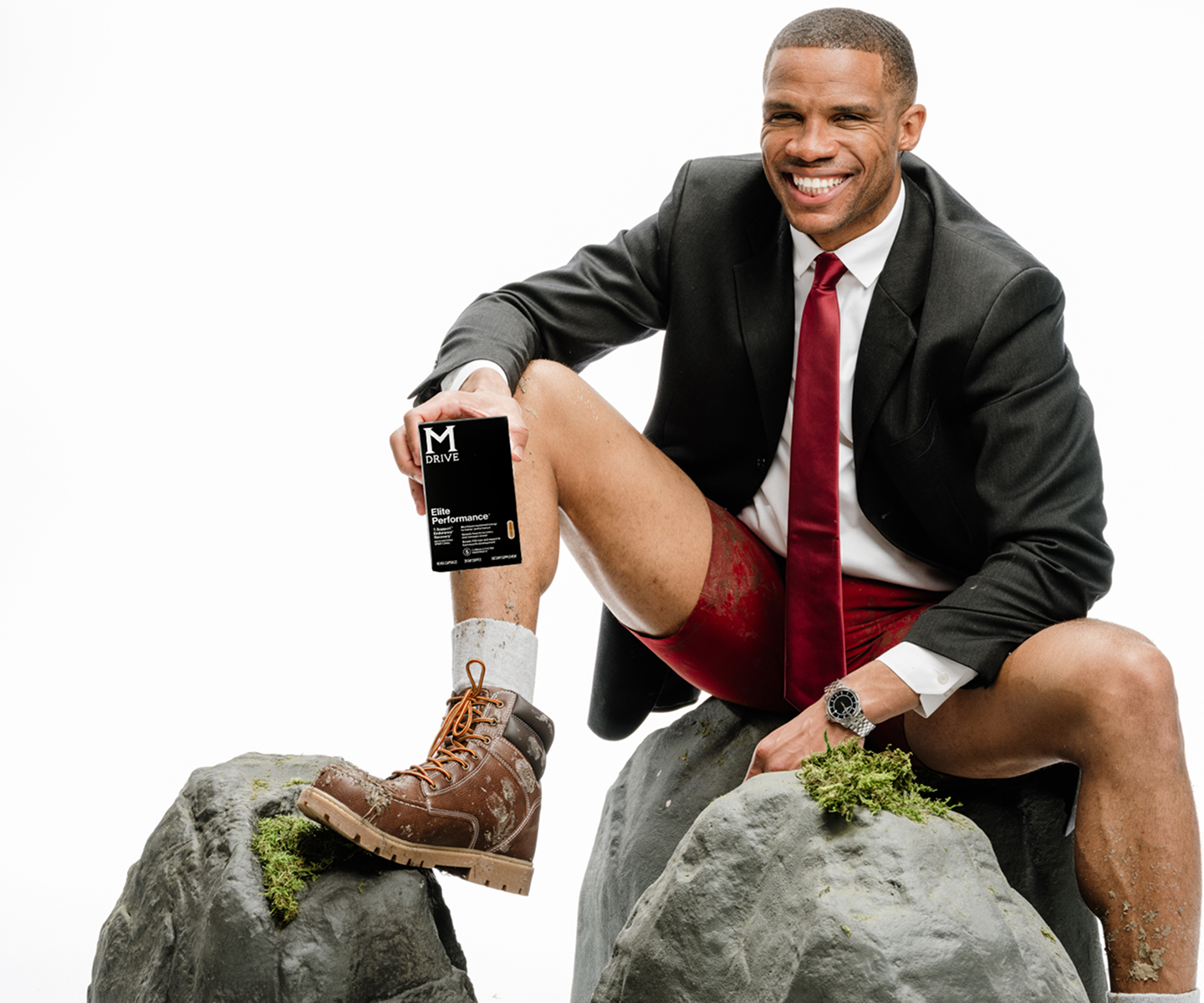Man in suit and boots sitting on rocks, holding a product box, smiling.