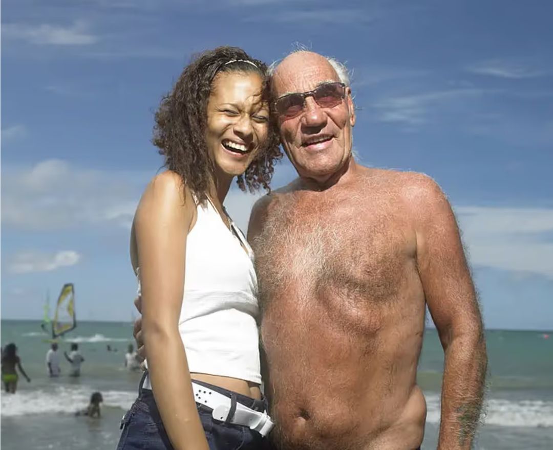 Two people smiling on a sunny beach with the sea and sky in the background.