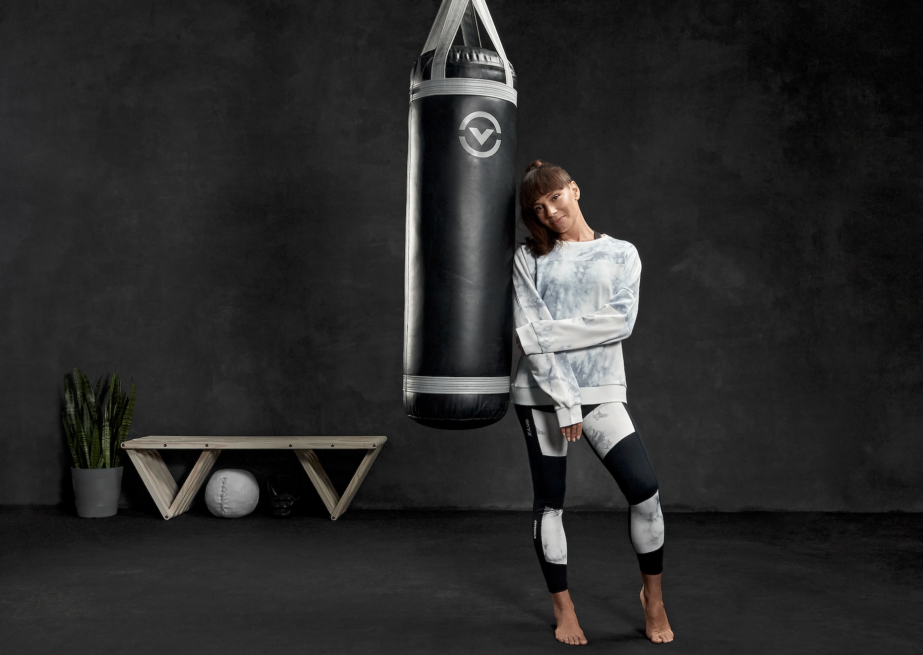 A woman in athletic wear leans against a black punching bag hanging in a dark room.