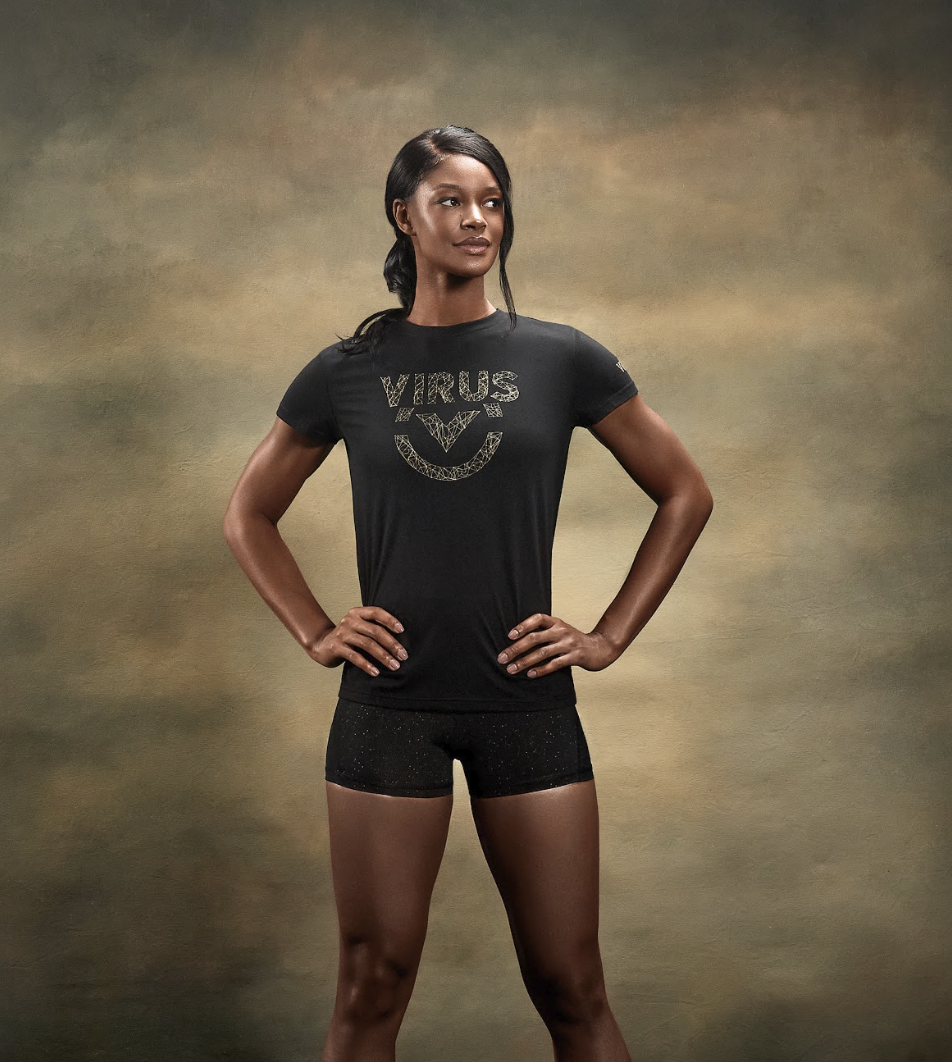 An athletic woman in a black VIRUS t-shirt and shorts poses with her hands on her hips.