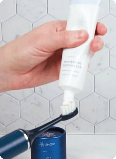 Hand applying toothpaste to an electric toothbrush in a bathroom setting.