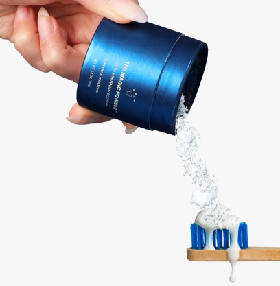 Hand pouring tooth powder onto toothbrush from a blue container.