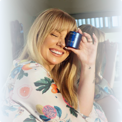Woman smiling, holding a blue cosmetic jar near her face, wearing a floral robe.