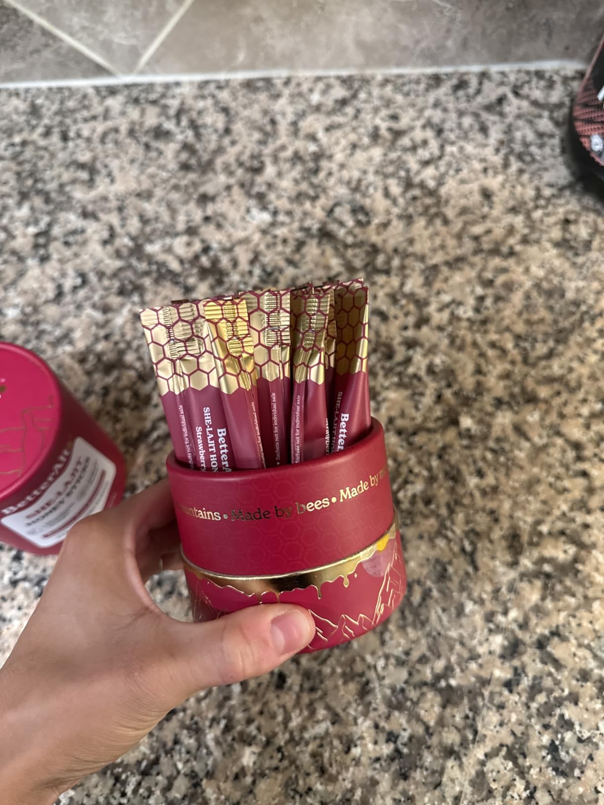Hand holding a circular container with red honey sticks on granite countertop.