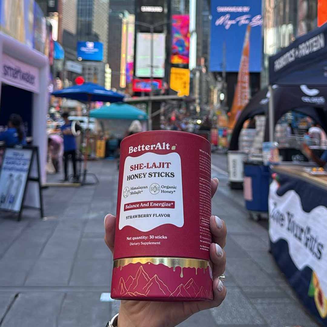 Hand holding a container of BetterAlt SHE-LAJIT honey sticks in a busy urban setting.