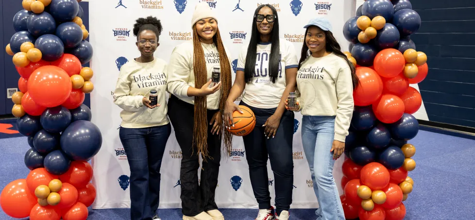 Four women pose with vitamin bottles and a basketball in front of a promotional backdrop with balloons.