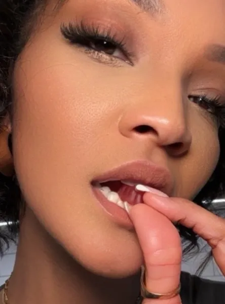 A close-up of a woman with makeup on, placing a finger in her mouth and touching her teeth.