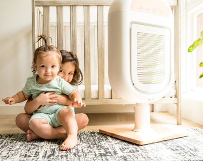Two young children sit on a rug in a nursery next to a white air purifier.