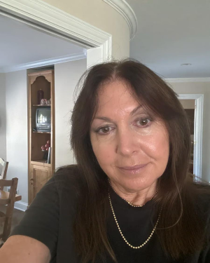 A woman with long dark hair and a silver necklace takes a selfie indoors.