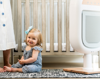 A smiling baby girl sits on a patterned rug in front of a wooden crib.