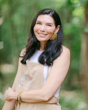 A smiling woman with dark hair, wearing a tan apron and a large earring, stands outdoors in a wooded area.
