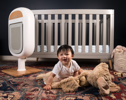 A smiling baby plays on a nursery floor next to a white air purifier and a crib.