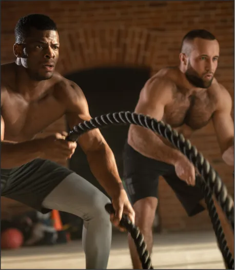 Two muscular men exercise with heavy battle ropes in a gym with brick walls.