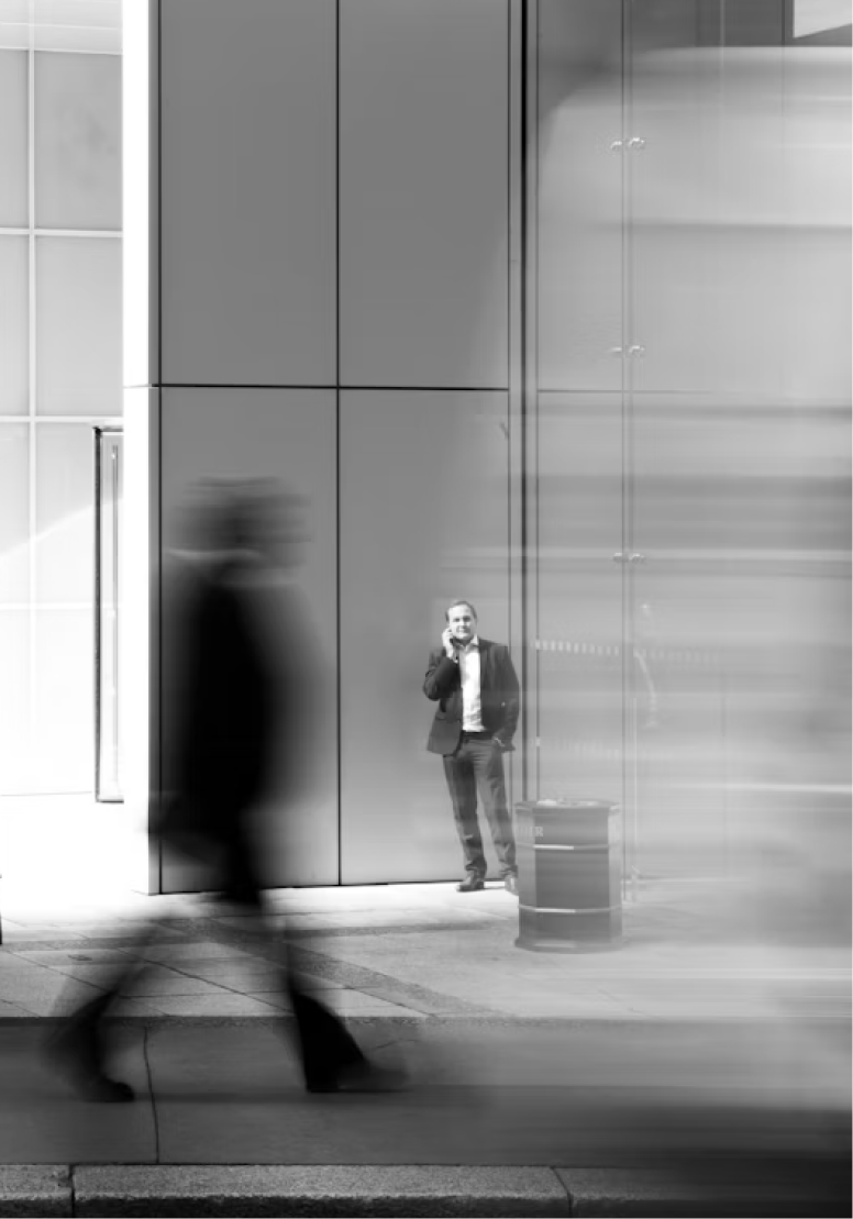 Man on phone stands against building, blurred pedestrians pass by.