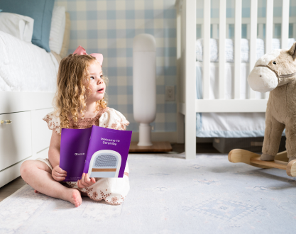 A young girl with curly hair sits on a bedroom floor holding a purple booklet.