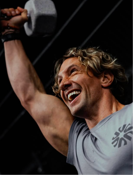 A smiling man with curly blond hair lifts a grey dumbbell over his head against a dark background.