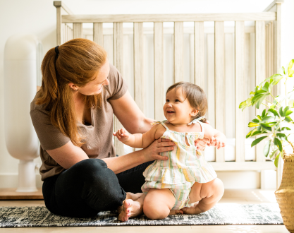 A woman sits on the floor holding a smiling baby in front of a crib.