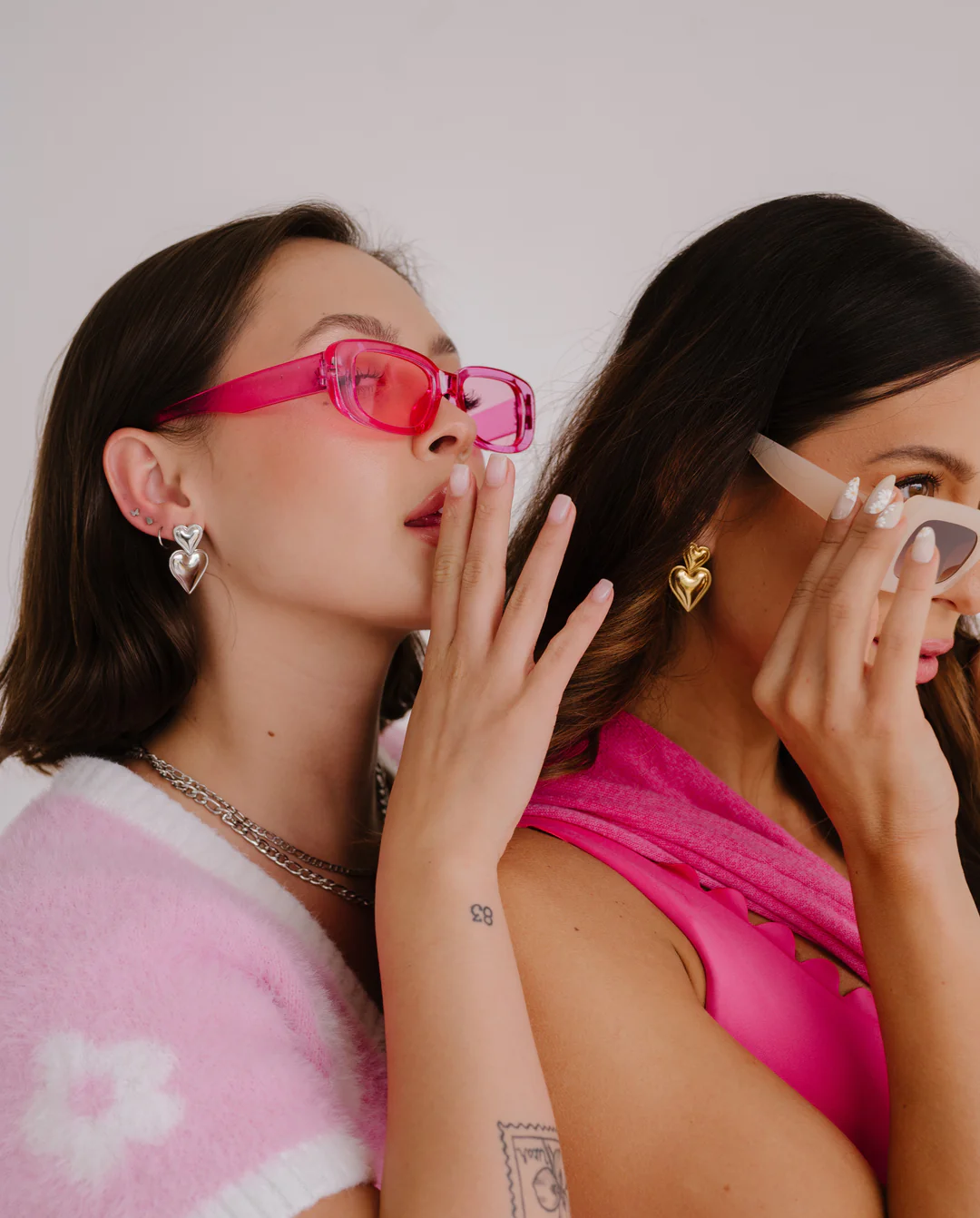 Two women wearing sunglasses and heart-shaped earrings, one whispering a secret to the other against a white background.