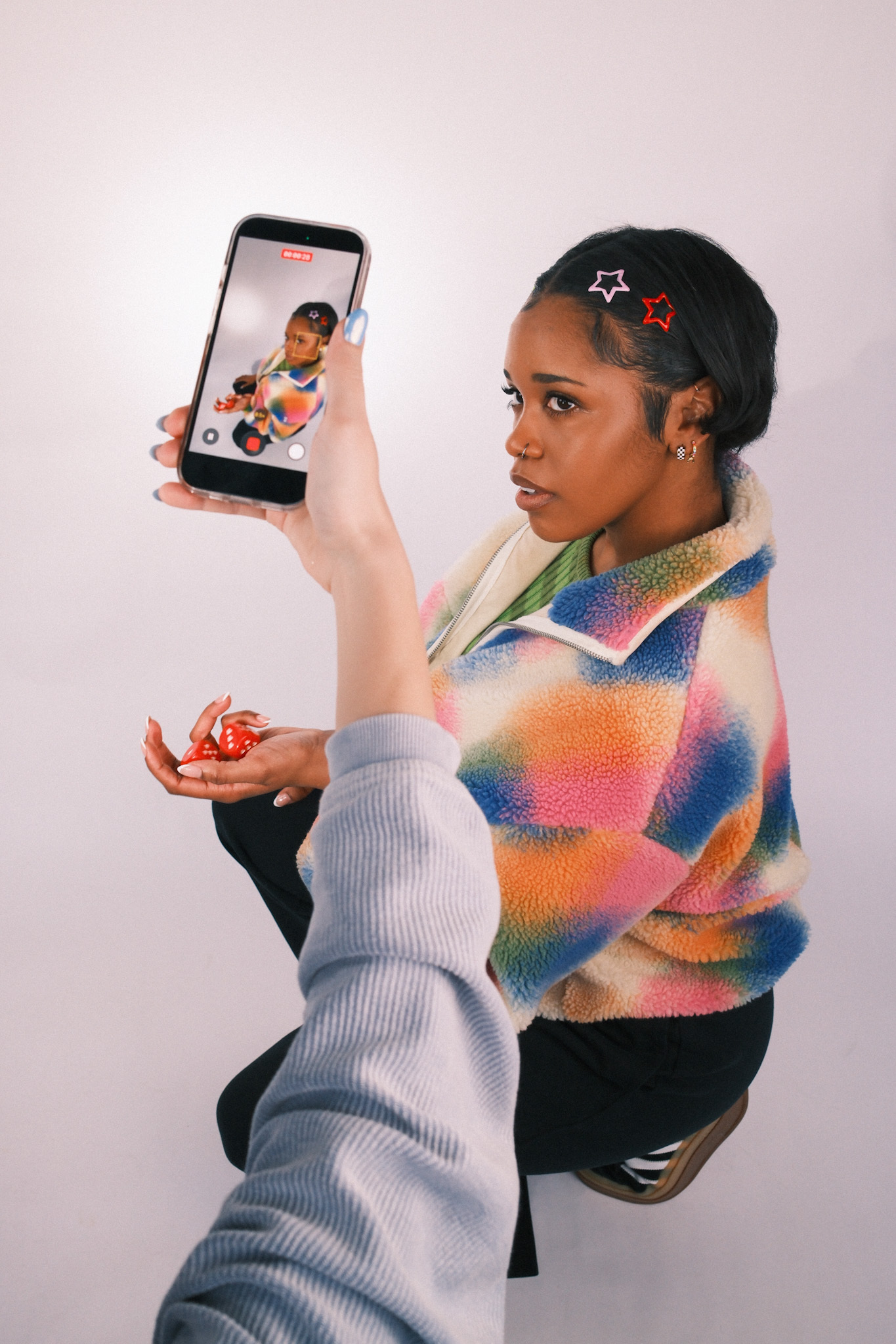 A person takes a smartphone video of a woman in a colorful fleece jacket holding red dice.