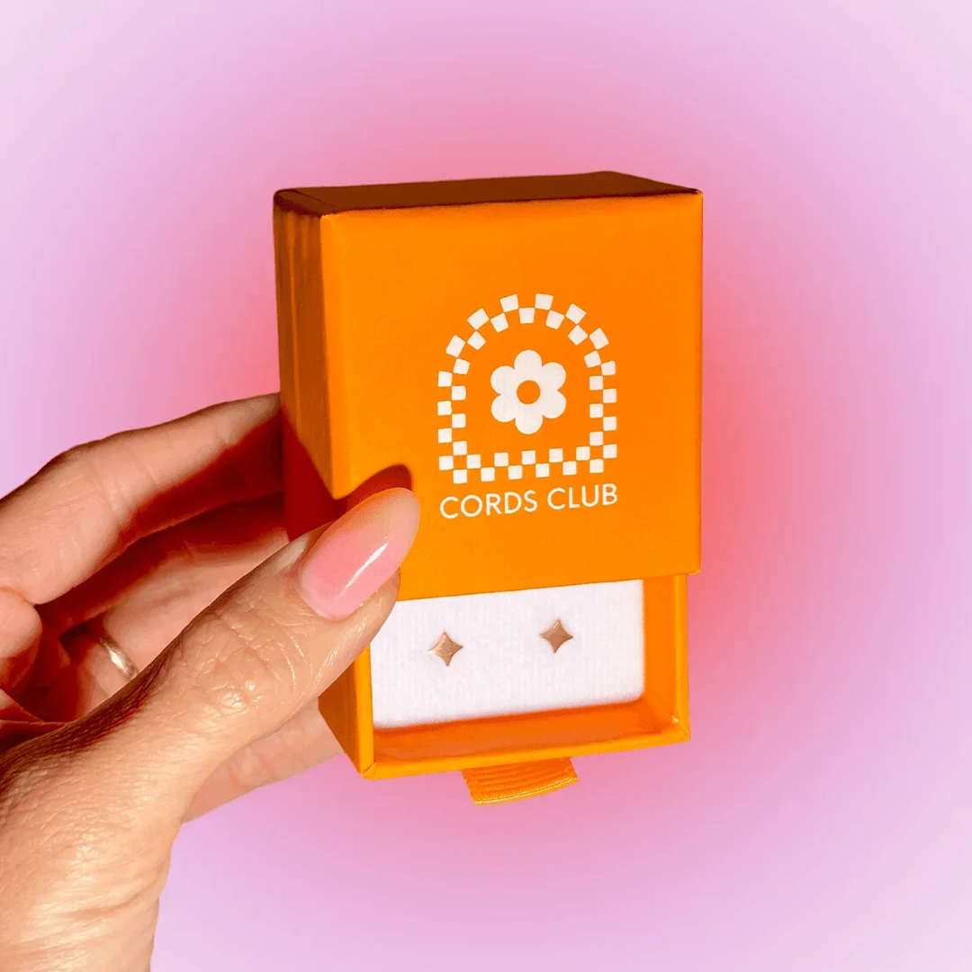 Hand holding an orange box labeled 'Cords Club' on a pink gradient background.