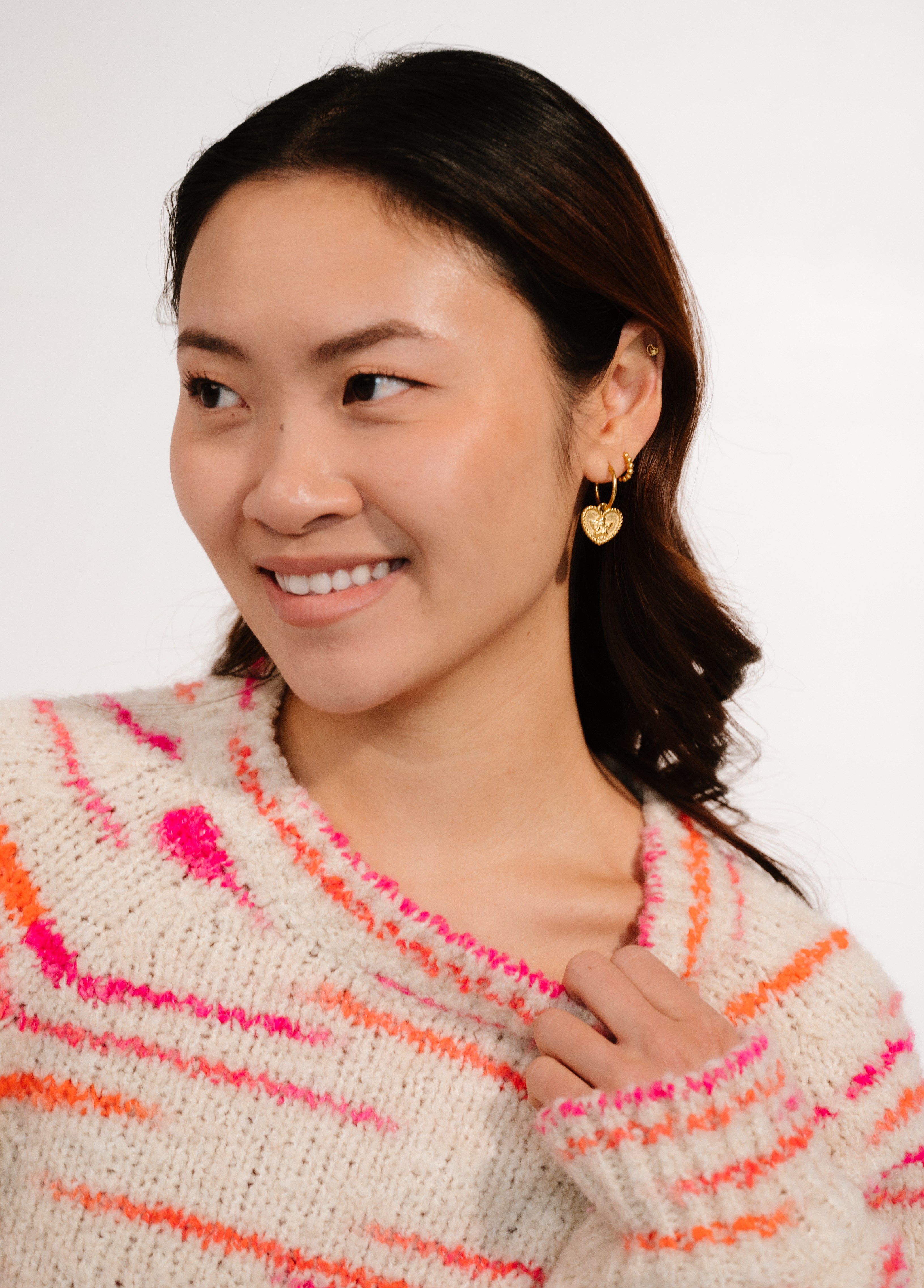 A smiling woman in a cream, pink, and orange striped sweater wears gold heart-shaped earrings.