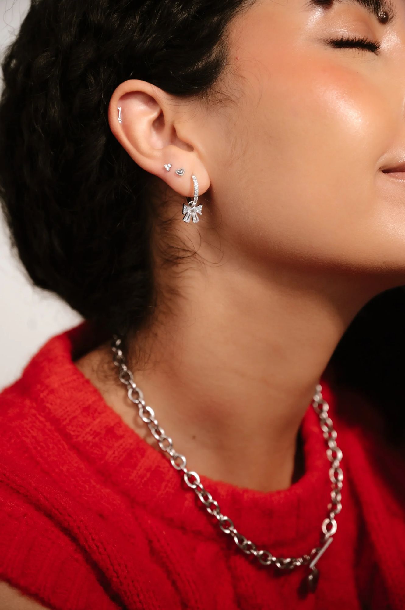 A close-up of a person in a red sweater wearing a silver chain necklace and multiple earrings.