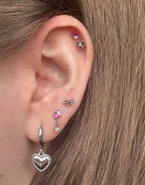 A close-up of an ear with multiple silver piercings, including heart, bow, and gem-shaped earrings.