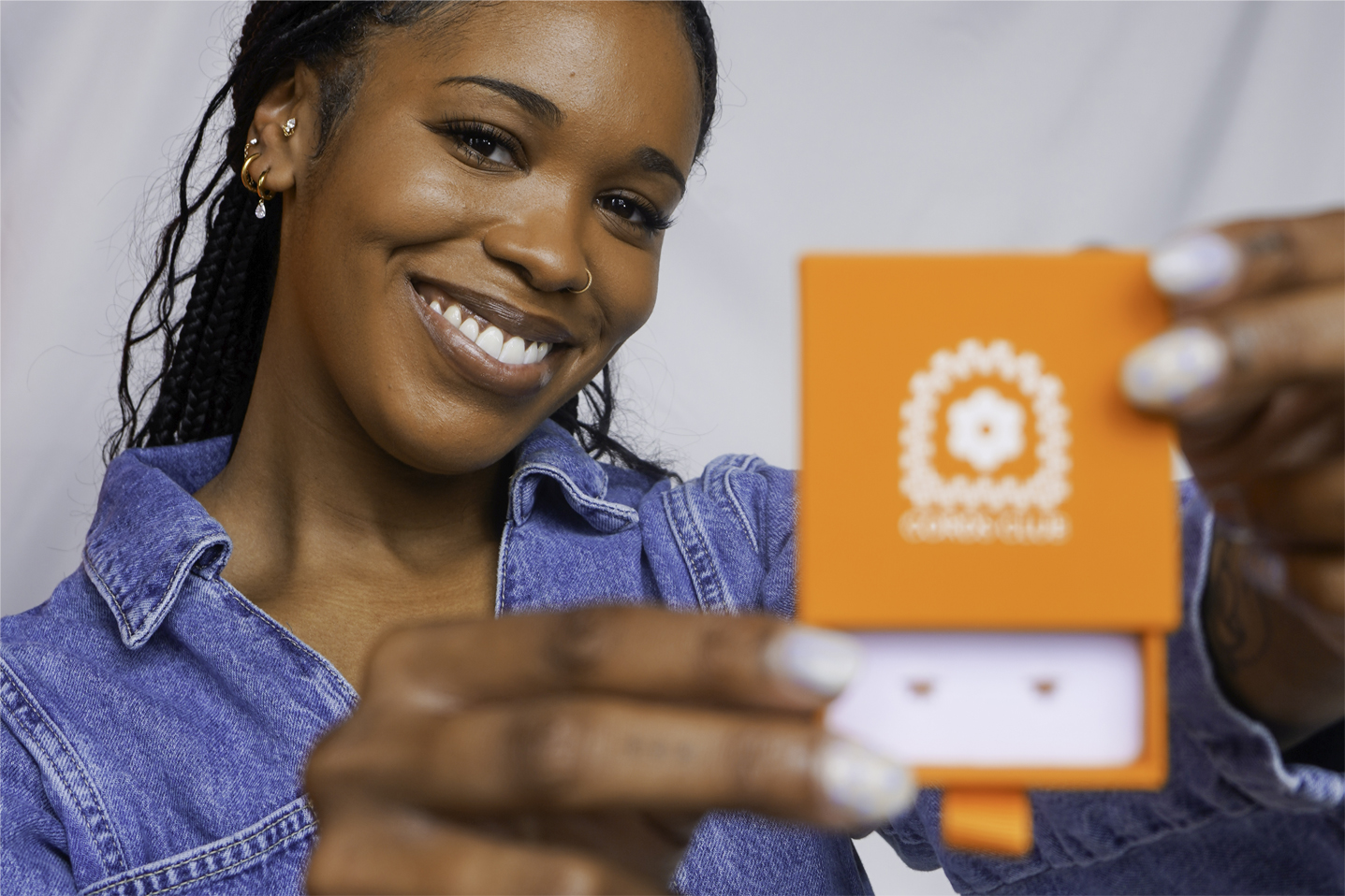 A smiling woman in a denim jacket holds an open orange jewelry box showing a pair of earrings.