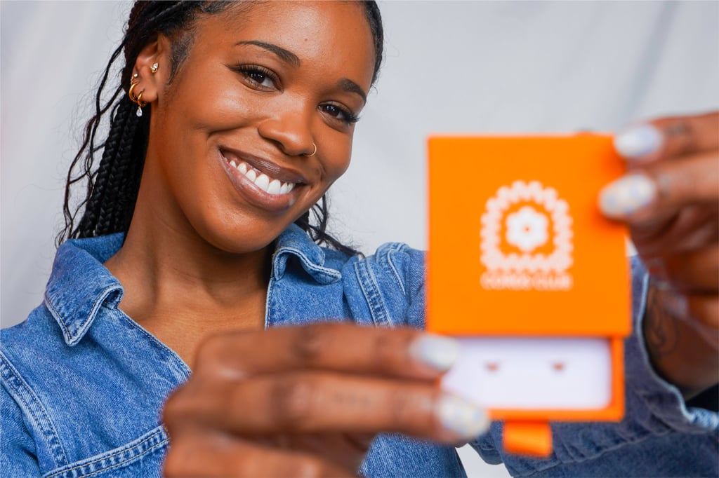 A smiling woman in a denim jacket holds an open orange jewelry box showing a pair of earrings.