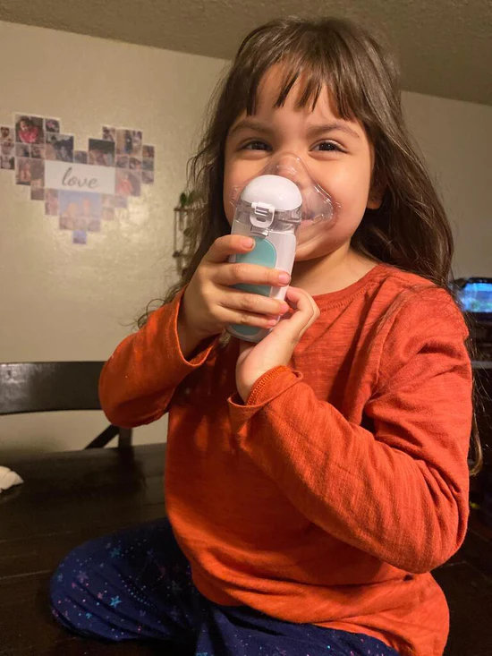 Child holding a nebulizer, wearing an orange shirt with a heart-shaped photo collage on the wall.