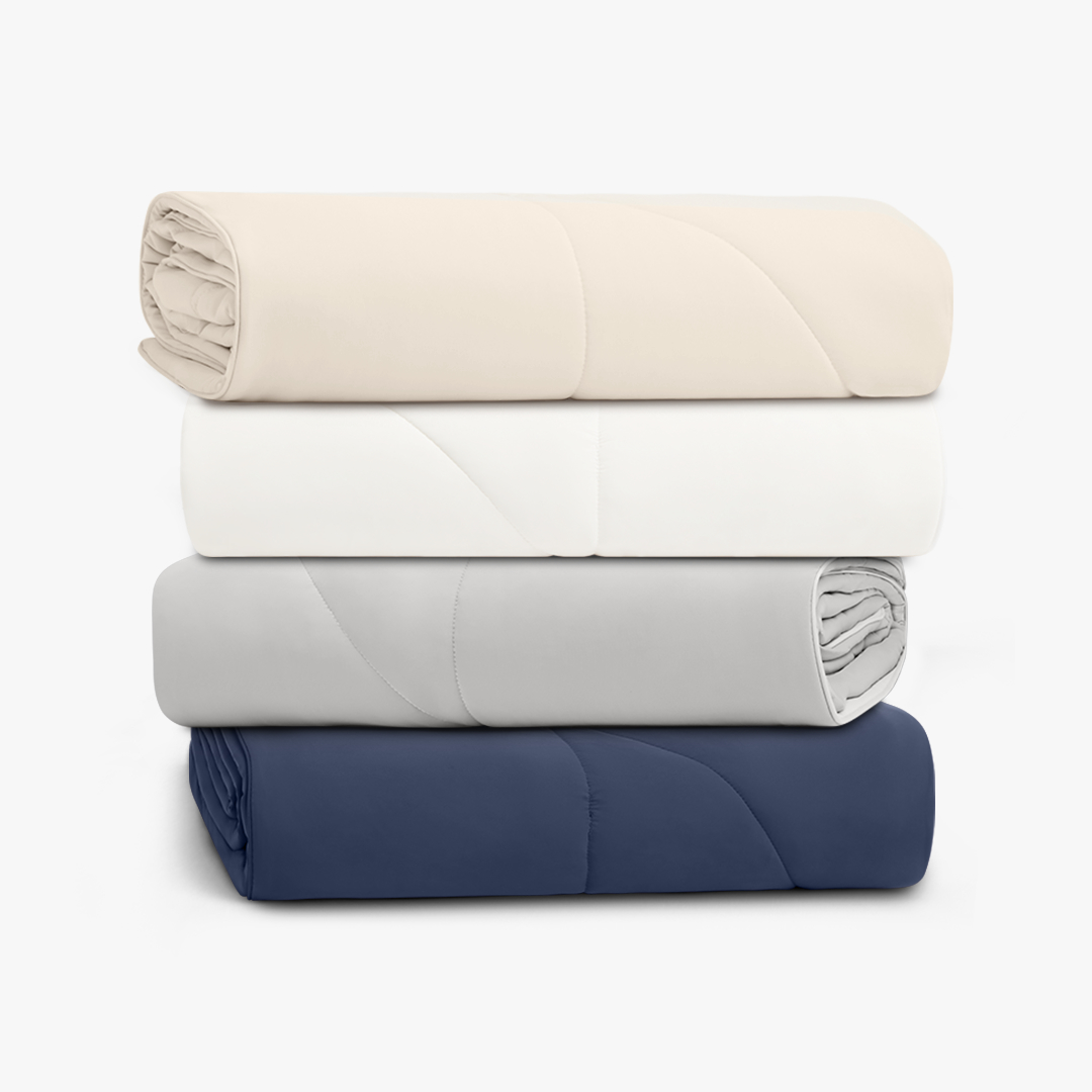 A stack of four neatly folded comforters in cream, white, gray, and navy blue against a white background.