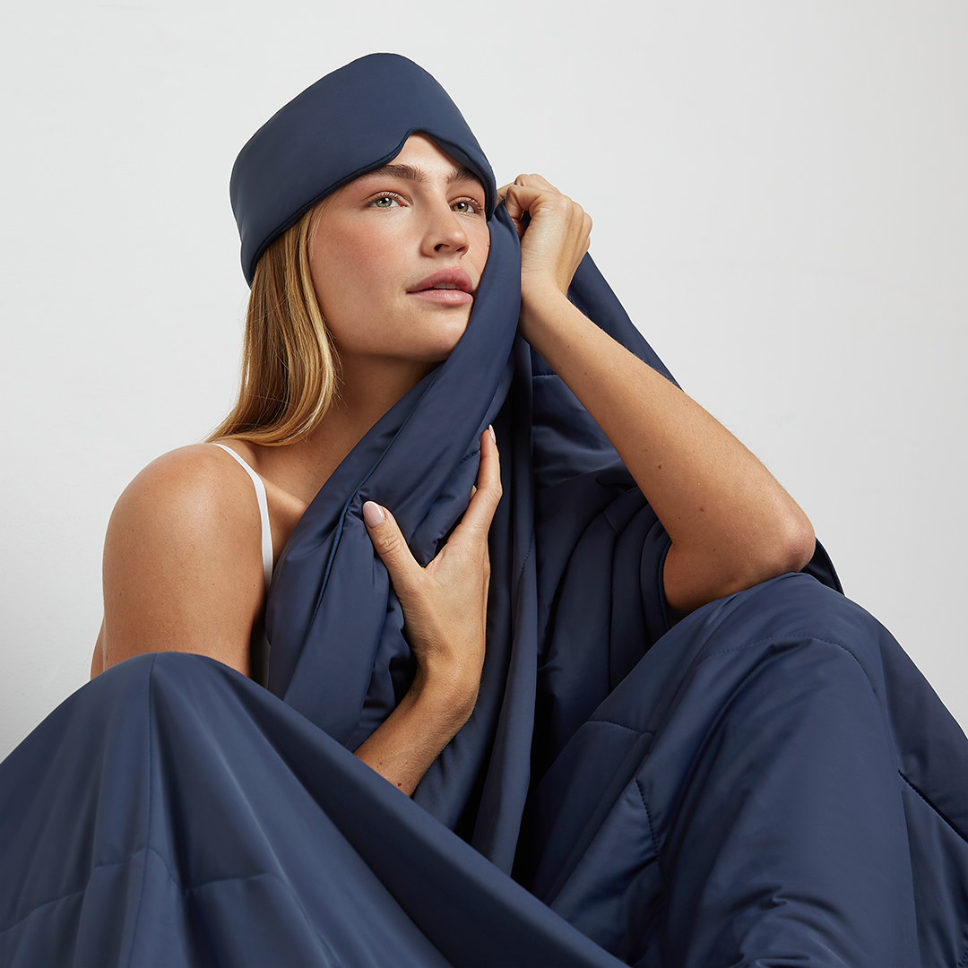 A woman wearing a blue sleep mask sits wrapped in a matching blue quilted comforter against a white background.