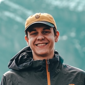 A young man in a yellow cap and dark hooded jacket smiles at the camera outdoors.