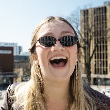 A young blonde woman in sunglasses laughs with her mouth open outdoors in a city.