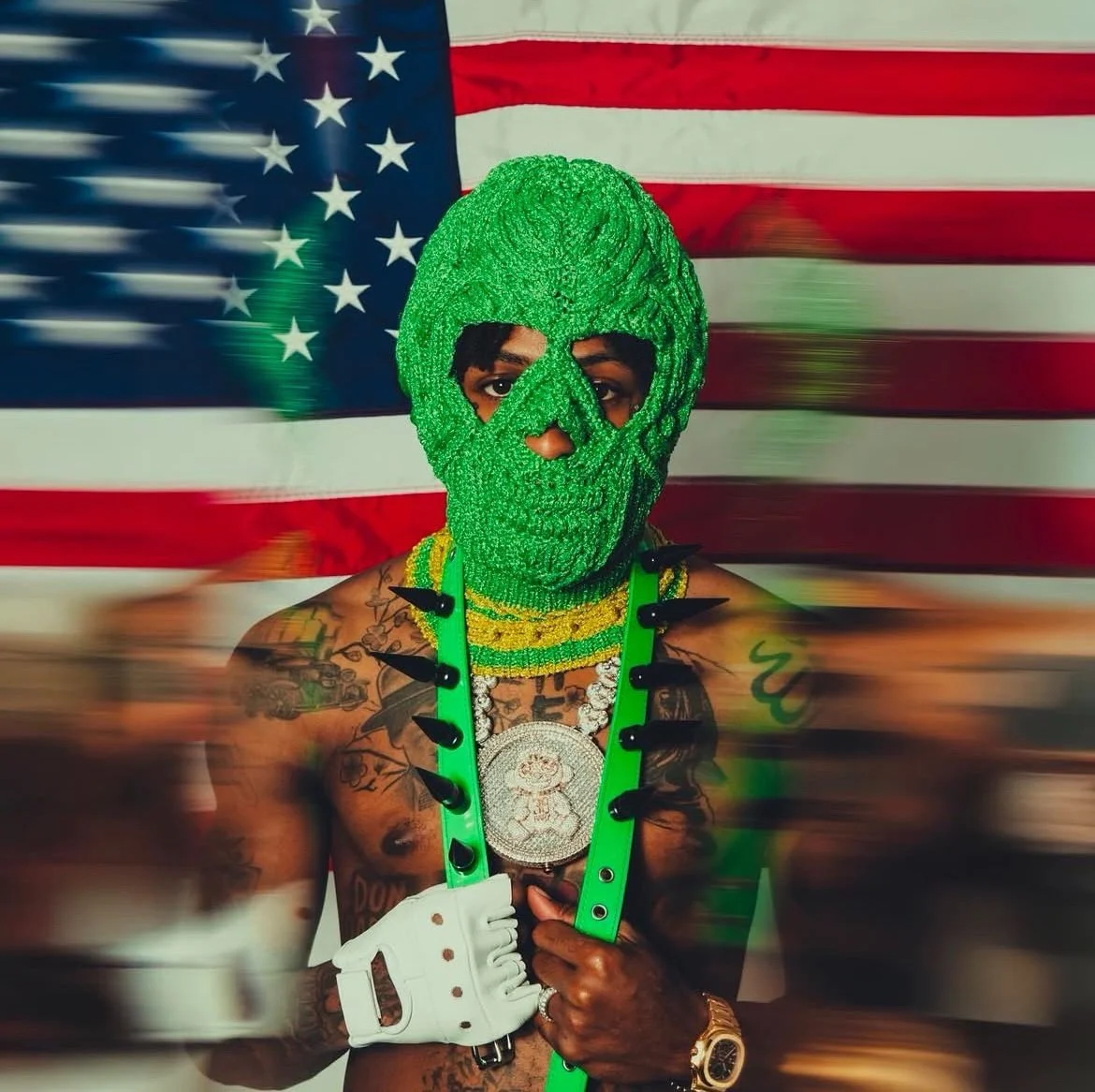 Person in green mask and accessories, standing in front of an American flag.