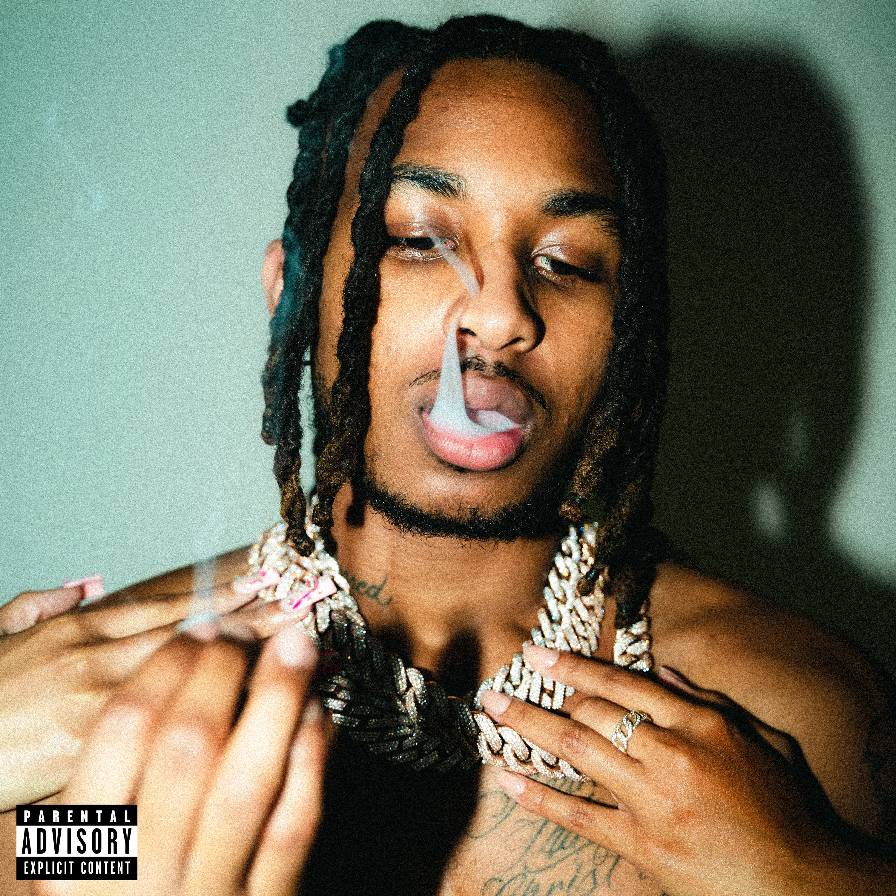 Person with dreadlocks and jewelry, exhaling smoke, against a plain background.