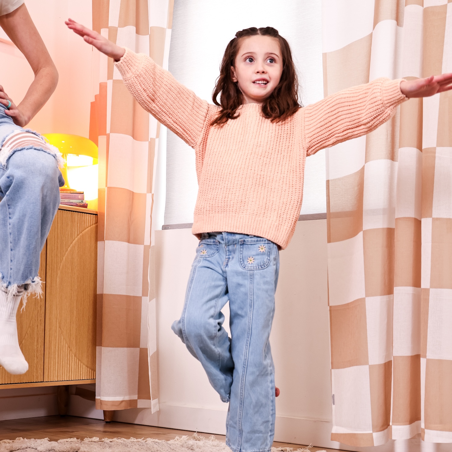 A young girl in a peach sweater and blue jeans balances on one leg with her arms outstretched.