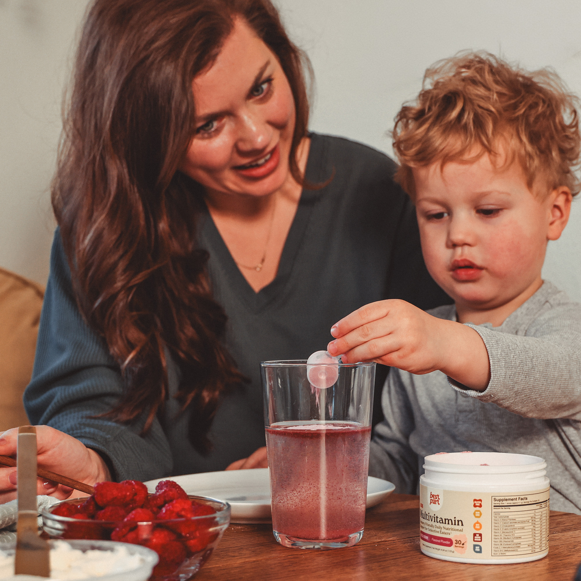 A mother watches her young son drop a multivitamin into a glass of water at a table.