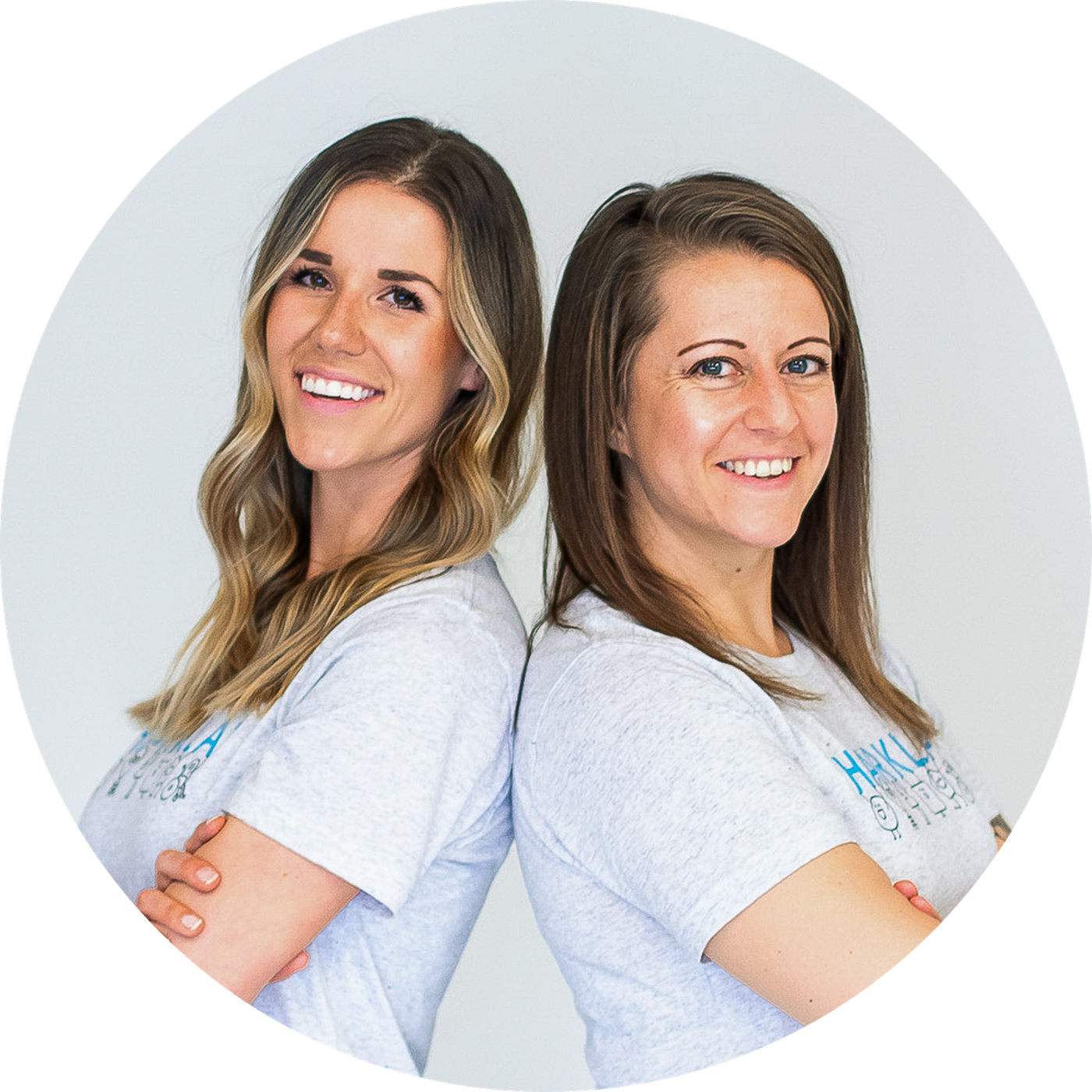 Two women stand back-to-back with their arms crossed, smiling at the camera in a circular portrait.