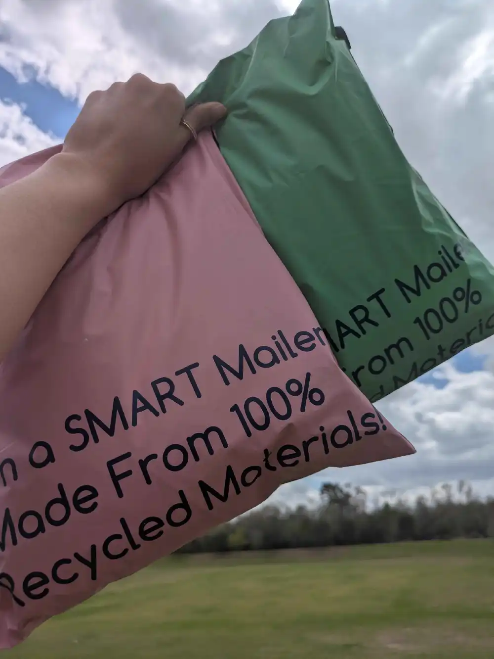 Pink and green recycled SMART mailers held outdoors in a grassy field representing sustainable shipping