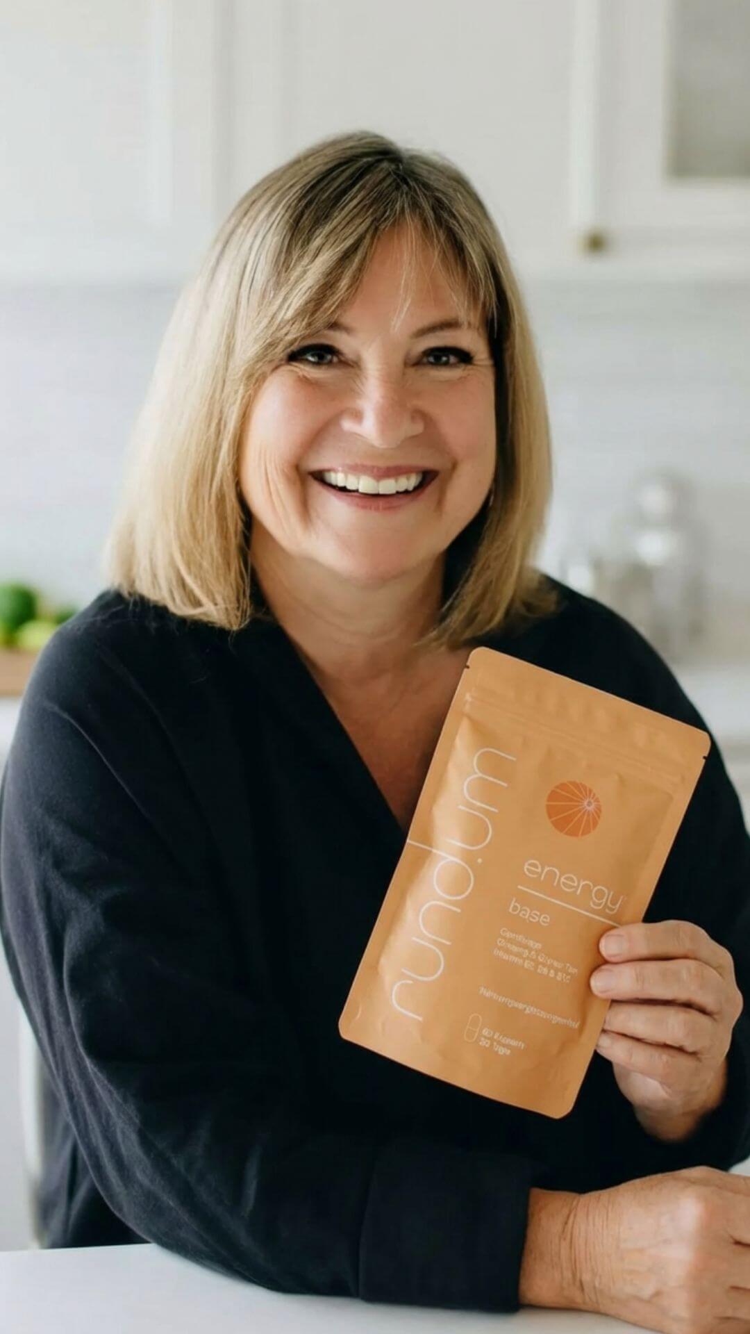 A smiling woman with blonde hair holds up an orange pouch of Rundum Energy Base supplement.