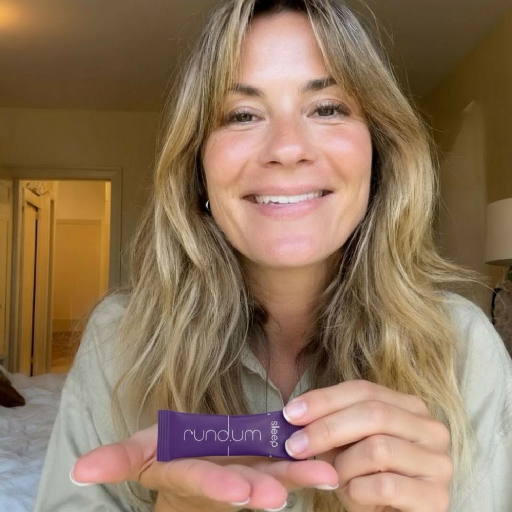 Person holding a small purple product labeled 'rundum sleep' in a bedroom setting.