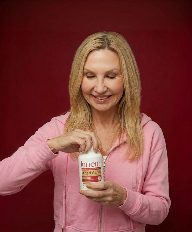 A smiling woman in a pink hoodie opens a white bottle of Lunera Aged Garlic against a red background.