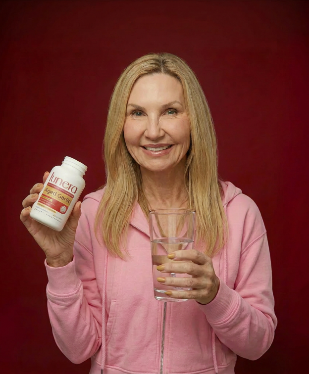 A smiling woman in a pink hoodie holds a bottle of Aged Garlic supplements and a glass of water.