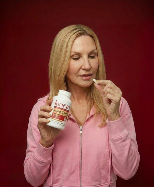 A woman in a pink hoodie holds a bottle of Lunera supplements and prepares to take a capsule.