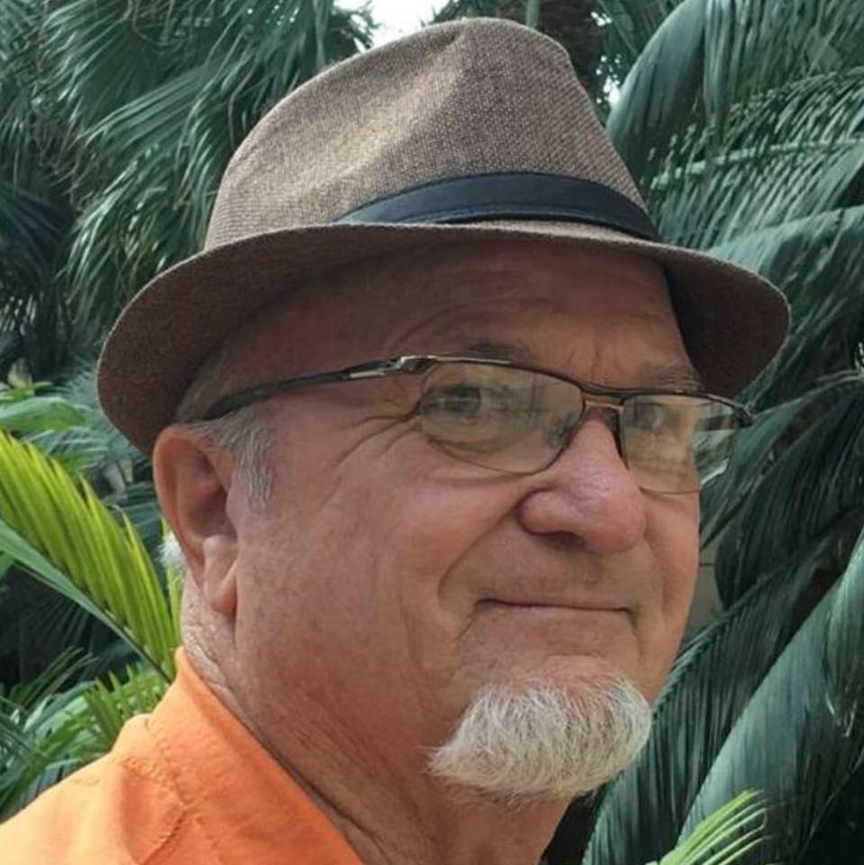 An older man with a goatee wearing a brown fedora and glasses, in front of palm leaves.