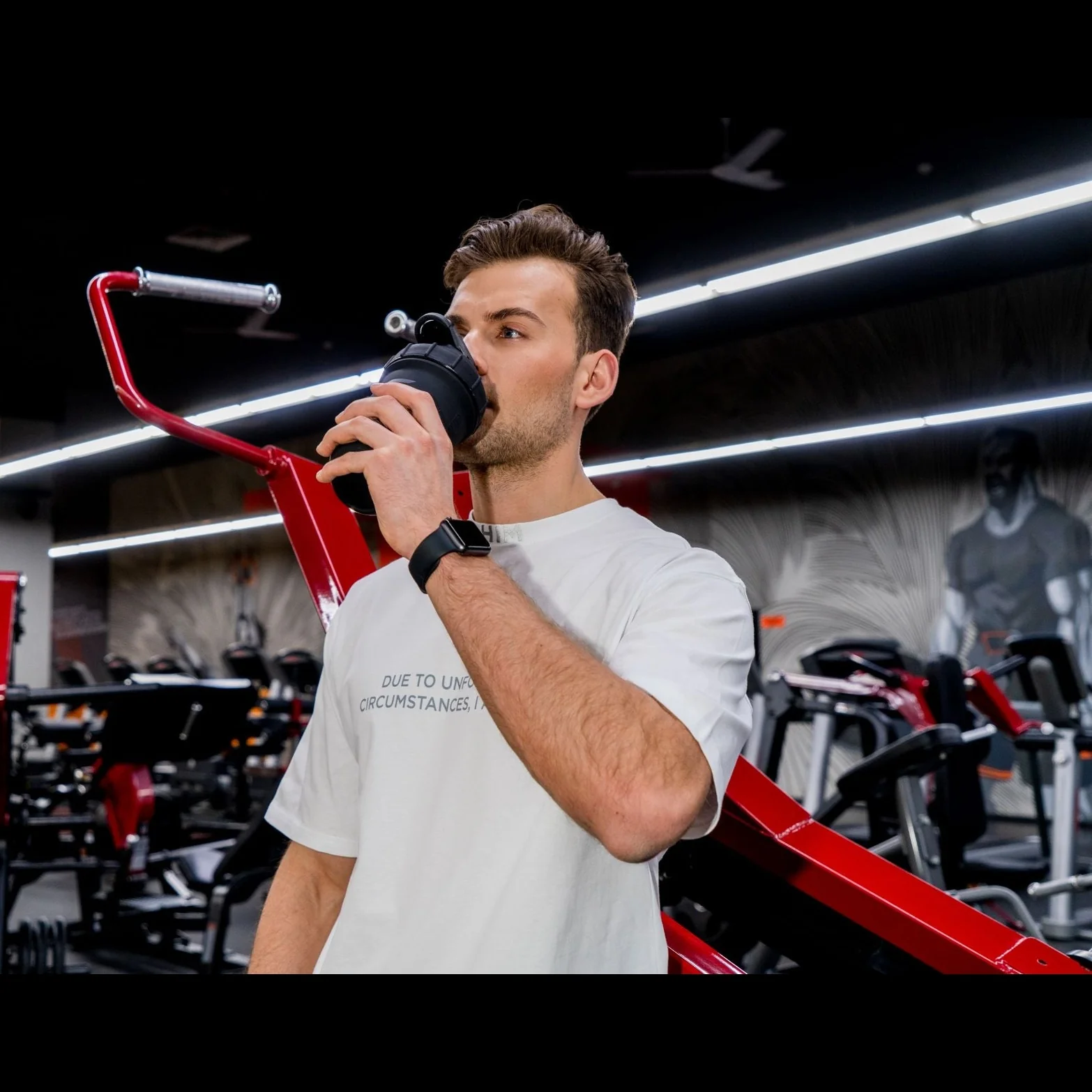 Man drinking from a shaker bottle in a gym with red equipment.